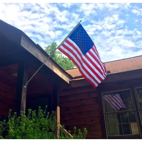 Nylon American Flag - Beacon® mounted on a wooden house exterior with green shrubbery and a partly cloudy sky background.