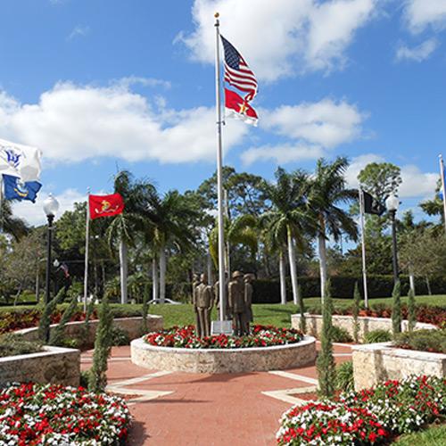 Nylon American Flag - Beacon® flying on a tall flagpole above a memorial with statues and surrounding tropical landscaping under a blue sky.