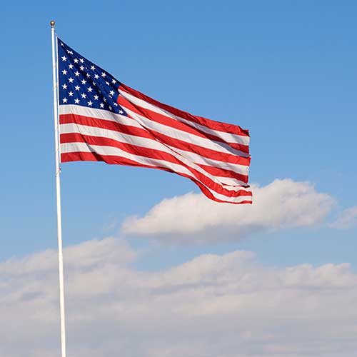 Nylon American Flag - Beacon® flying on a flagpole against a clear blue sky with light clouds, showing vibrant red, white, and blue colors.