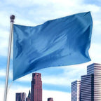Blue flag waving on a pole against a city skyline, made of durable material in Solid Colored Flags - Premier Nylon.