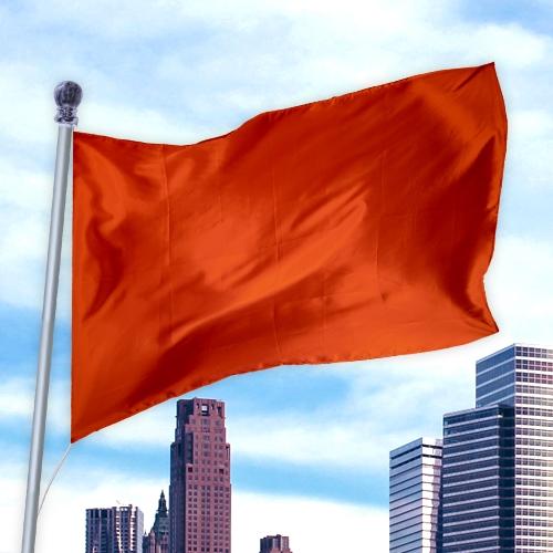 Solid Colored Flags - Premier Nylon in bright orange waving outdoors against a blue sky and city skyline background