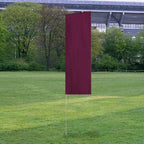 Solid Tall Advertising Flags in a deep red color standing upright on a green grassy field with trees and a stadium in the background.