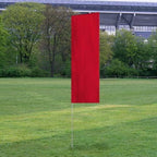 Solid Tall Advertising Flags in a bright red color standing upright on a grassy field with trees and a stadium in the background.