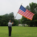 Man in uniform holding a Jointed Oak Parade Pole Set With U.S. Flag outdoors on a grassy field with trees in the background.