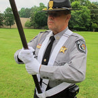 Man in sheriff uniform holding the Jointed Oak Parade Pole Set With U.S. Flag during an outdoor ceremony on a grassy field.